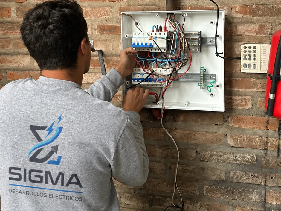 Técnico de Sigma, de espaldas con el logo en el buzo, conectando un tablero eléctrico sobre una pared de ladrillo
